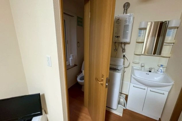 寮の洗面・洗濯スペースと、その奥に見えるトイレのドア。手前に小型テレビと洗面台があり、壁には給湯器と洗濯機が設置されている。