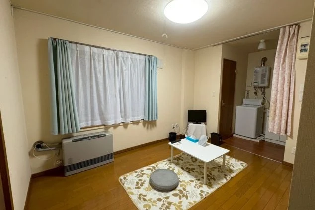 寮の共有リビングまたは居室。窓際に大型のFF式ストーブがあり、中央にはローテーブルとラグが敷かれている。奥のスペースには給湯器と洗濯機が見える。