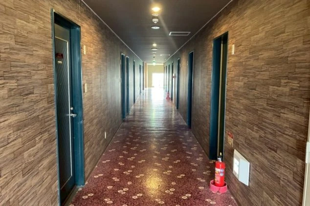 寮の居住フロアの長い廊下。両側に扉が並び、奥は明るい。壁と床は落ち着いた木目調。