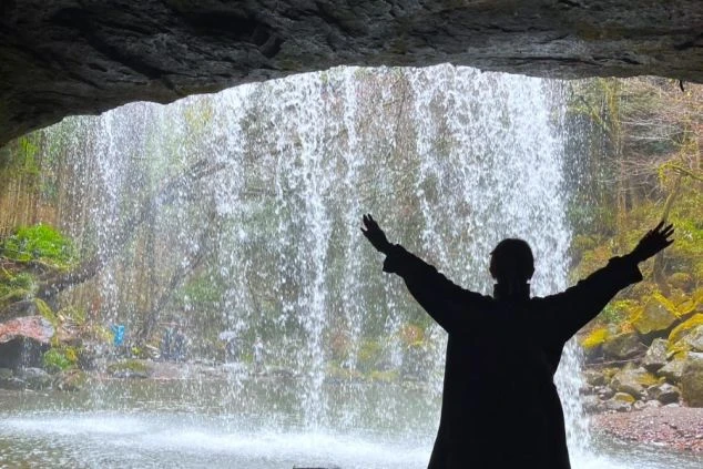 熊本県小国町にある「鍋ヶ滝」の裏側から、降り注ぐ水のカーテンを眺め、両手を広げるリゾートバイトスタッフのシルエット。阿蘇エリアのホテルでの住み込みバイト、休日の観光イメージ。