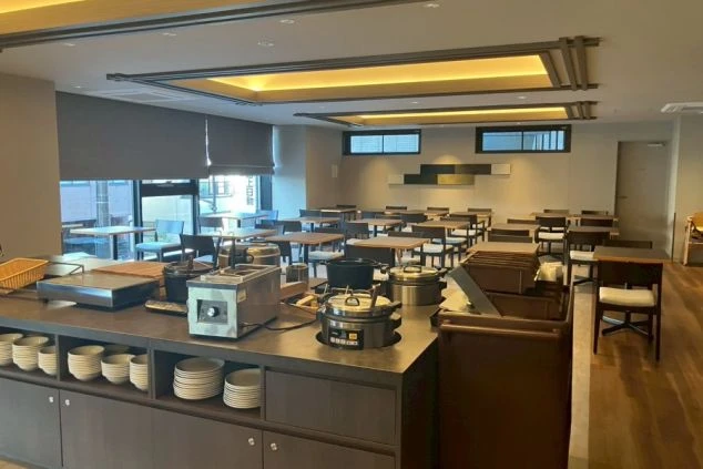 レストラン（ビュッフェ会場）。ビュッフェカウンターに料理の保温容器などが置かれ、奥には木製テーブルと椅子が規則的に並んでいる。