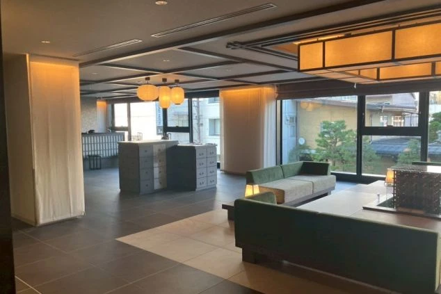 ロビーまたはラウンジ。モダンな内装で、ダークトーンの床、緑のソファ、和風の間接照明があり、奥にはチェックインカウンターと思われるスペースが見える。