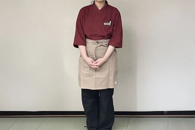 赤茶色の七分袖の作務衣とベージュのエプロンを着用した女性が、正面を向いて立っている様子。