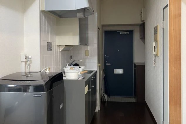 玄関と廊下が写されていて中央にはレンジフードのあるコンロとシンクエリアが見えます。カウンタートップは暗い色で、その下には引き出しや戸棚があるように見えます。