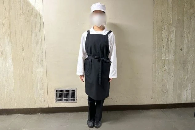 白い調理服に黒のエプロン、マスクを着用し帽子をかぶった従業員食堂の調理補助の制服画像。
