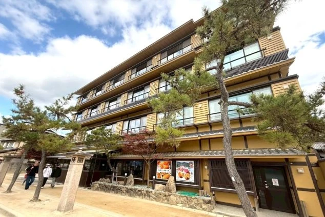 松の木に囲まれた、瓦屋根と茶色い壁が特徴の日本的な外観を持つ、数階建ての宿泊施設。