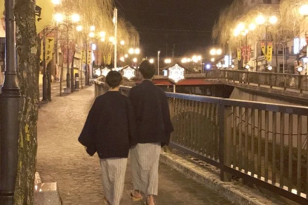 リゾートバイト後の温泉街を浴衣で夜に散策する二人組の画像。柳や提灯の明かりが情緒ある温泉地の風景。