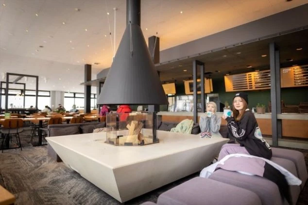 ゲレンデのレストハウスやロビー。中央にはモダンなデザインの暖炉があり、ソファでくつろぐ2人の女性が飲み物を飲んでいる。