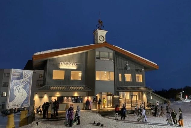 夜間、雪の上に建つ、時計台のような屋根を持つ洒落た建物。窓から光が漏れ、人々がチケットカウンターの周辺に集まっている。