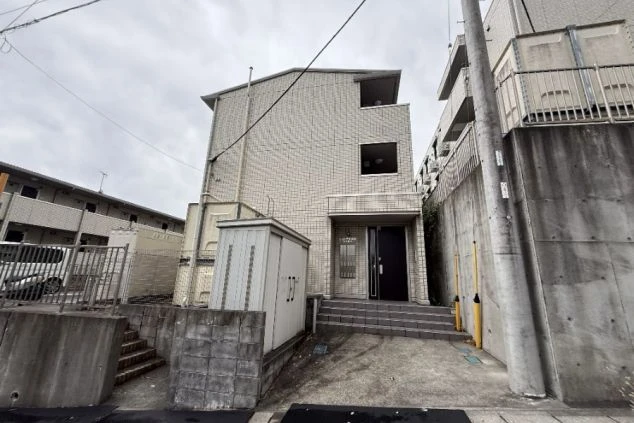 寮の外観。灰色のタイル壁の三階建てほどの建物で、周囲はコンクリート壁に囲まれている。