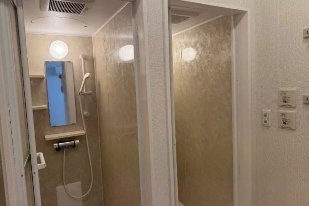 共有のシャワールームまたは個室のユニットバス内。ベージュのパネル壁で、シャワーヘッドと鏡が設置されている。