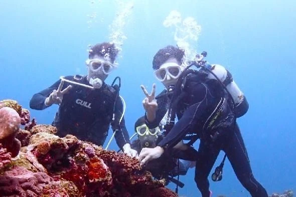 明度の高い海中で、ダイビングスーツを着た3人が水中カメラに向かってピースサインをしている。手前には鮮やかなサンゴが見える。
