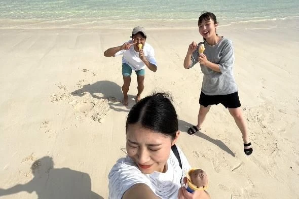 白い砂浜で、アイスクリームを持った若い男女3人が自撮りをしている。手前の女性がカメラを構え、奥の2人はピースサインをしている。