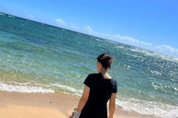 快晴のビーチで、黒いTシャツを着た女性が海を背にして立っている。青い海と白い波が輝き、リゾート地でのリラックスした様子。