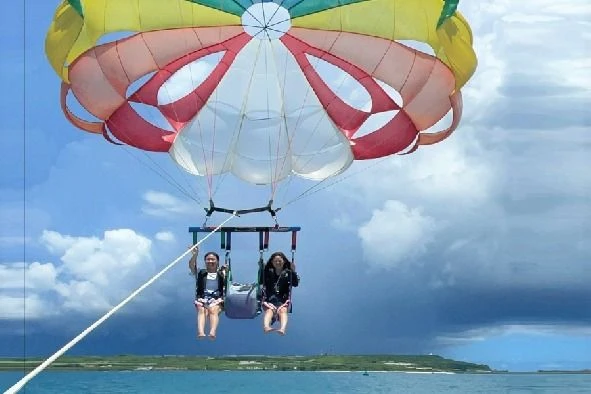 青空の下、海の上空でパラセーリングを楽しむ若い男女2人組。色鮮やかなパラシュートに吊り下げられ、海と島を眺めている。
