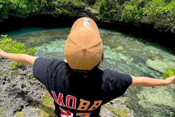 茶色のキャップと黒いTシャツ姿の人物が、両手を広げて岩場の上に立っている。眼下には透明度の高い青い水面が広がる洞窟のような場所。