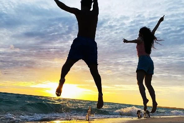 夕焼け前のビーチで、男女が大きくジャンプしているシルエット写真。海辺で手足を広げ、ダイナミックに楽しんでいる様子。