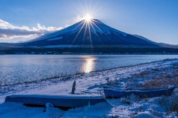 冬の早朝、雪をかぶった富士山頂から太陽の光が差し込み、湖面に反射している雄大な風景。