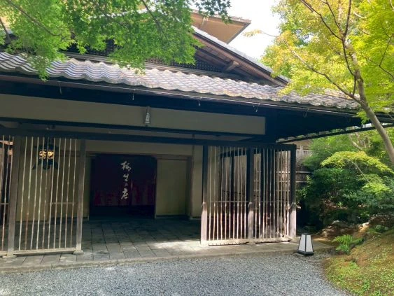 伝統的な日本の旅館の玄関。瓦屋根のついた重厚な門構えで、木製の縦格子戸が開いている。玄関奥には「梅の屋」などと読める文字が書かれた赤い暖簾が見え、周りは青々とした木々に囲まれている。