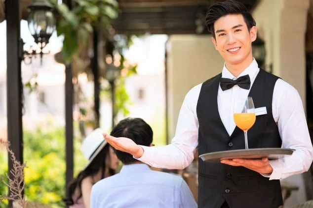 レストランの男性ウェイターがドリンクを片手にお客様を案内する様子。レストランのリゾートバイトの業務イメージ