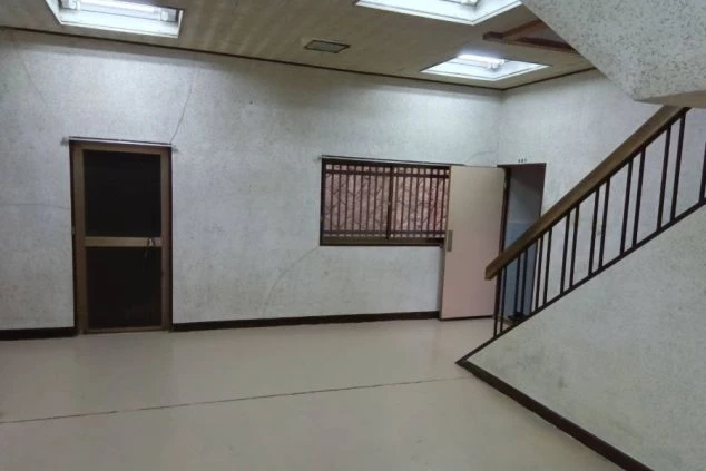 木目調の壁と白いタイル床の部屋に、シングルベッドとエアコン、窓の外に瓦屋根が見える。