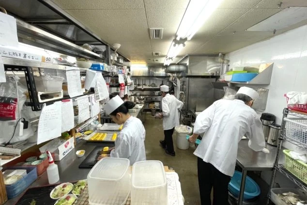 白いコックコート姿の複数人の料理人が、雑然とした厨房内で調理作業をしている様子。