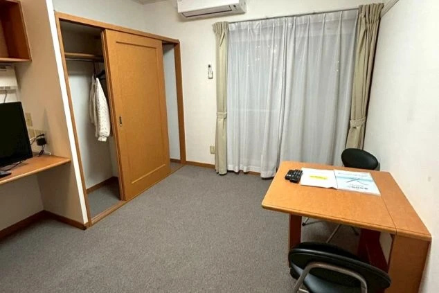 従業員寮の個室(居室)。カーペット敷きの床に、窓からの明るい光が差し込み、奥にはクローゼットと机が見える。