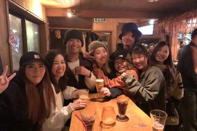 バーのような屋内で、ビールや飲み物を持った数人の若者が、テーブルを囲んで笑顔で集まっている。