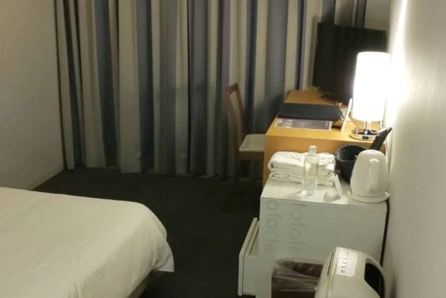 従業員寮の個室。ベッド、デスク、テレビ、電気ポット、ミネラルウォーターが置かれ、窓には青と白の縦縞のカーテンがかかっている。