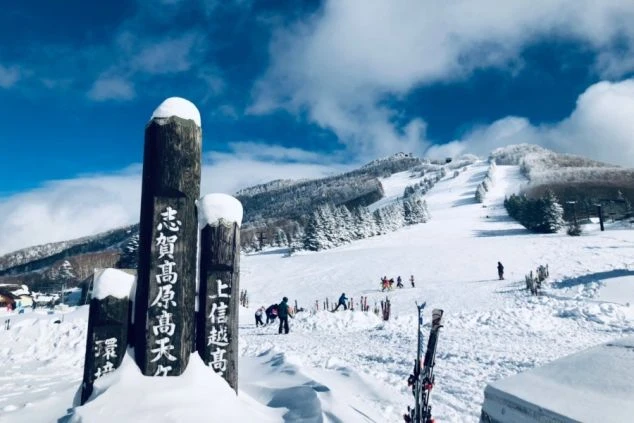 雪が積もった道標（どうひょう）と、賑わう志賀高原のスキー場の晴れた日の景色の画像。