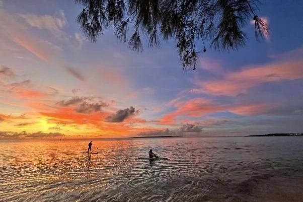 鮮やかなオレンジ色に染まる空と海を背景に、SUP（スタンドアップパドルボード）を楽しむ人々のシルエット。リゾートバイトの仕事終わりや休日の過ごし方イメージ。