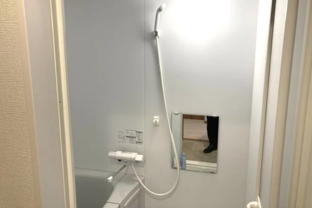 白い壁の浴室。左奥に浴槽があり、中央にシャワーと小さな鏡が設置されている。