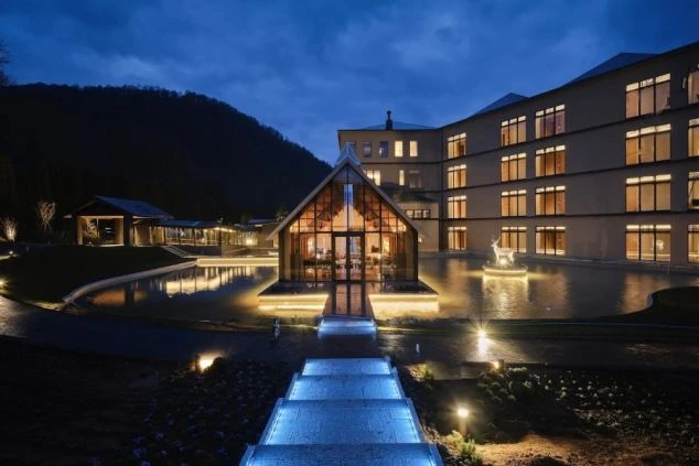 夜、水辺にガラス張りの小さな建物と、複数の窓に灯りがともる低層のモダンなホテル。