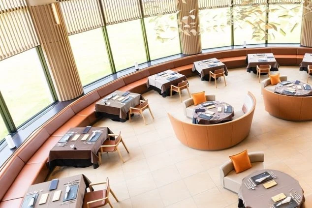 丸い窓から外の緑が見える、カーブしたソファと丸テーブルが配置されたモダンなレストラン。