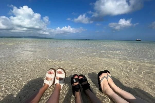 ビーチでリゾートバイト仲間とリラックスする足元のカット。白い砂浜と透き通る浅瀬の海を背景に、サンダル姿で並ぶ友人同士。住み込みバイトで出会う新しい仲間との思い出。