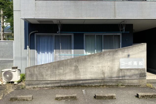 グレーのタイル外壁の建物の一角。手前にはコンクリートの低い塀があり、駐車場が隣接している。