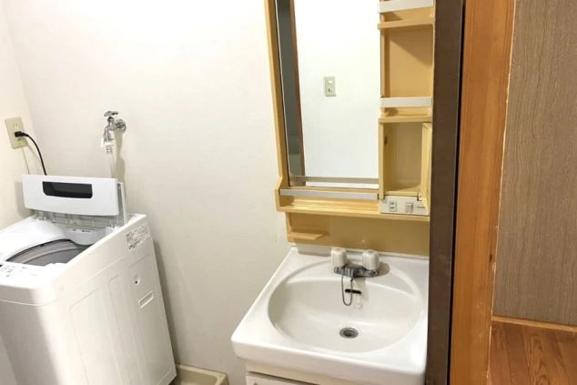 洗面脱衣所。白い洗面台の横に縦型洗濯機が置かれ、壁には鏡付きの収納ラックが設置されている。