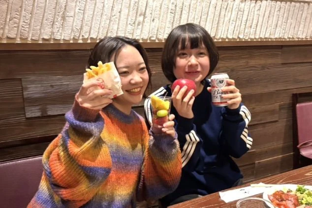 居酒屋の席で、フライドポテトやリンゴ、缶ビールを手に持ち、笑顔で楽しそうに過ごす2人の女性。
