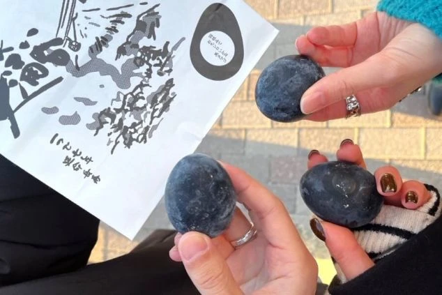 3人の人物がそれぞれ手に持った、真っ黒な殻のゆで卵（黒たまご）。背景には商品の袋が見える。