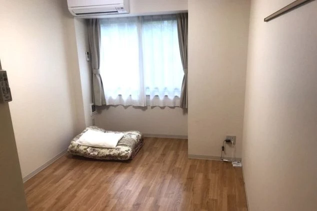 寮の居室。フローリングの床で、窓際に布団が敷かれている。部屋にはエアコンが設置され、テレビ台と小型テレビが見える。