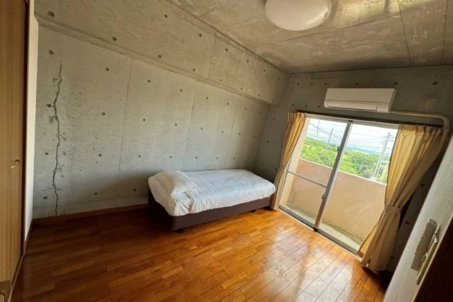 シンプルなベッドルーム。窓から光が差し込み、海の景色が見える。部屋にはベッドとエアコンがある。