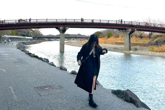 川辺の遊歩道を歩き、腕時計を確認する女性の姿。リゾートバイトの休日に、伊勢の風情ある街並みを散策するイメージ。