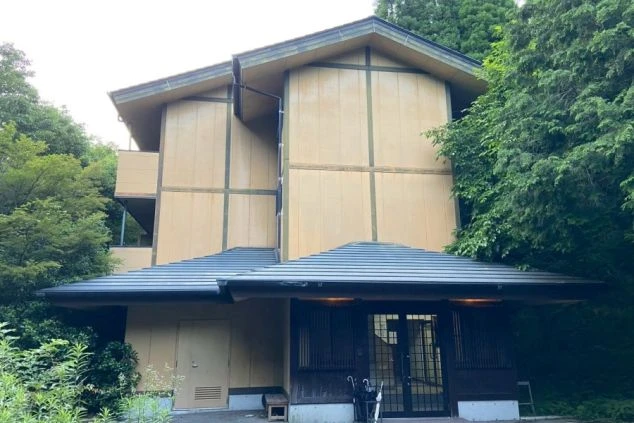 木々に囲まれた場所に建つ、和風の屋根とベージュの外壁が特徴的な2階建て住宅風の建物。