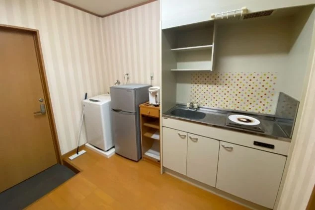 室内に洗濯機、冷蔵庫、電子レンジ、一口コンロ付きのキッチンがコンパクトにまとまった生活スペース。