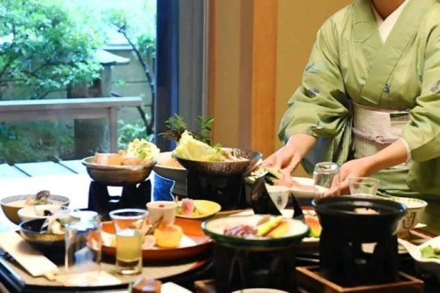 若草色の着物を身にまとった仲居スタッフが、彩り豊かな懐石料理を丁寧に配膳する様子。本格的な和食マナーやおもてなしを学べる、高級旅館でのリゾートバイト業務イメージ画像。