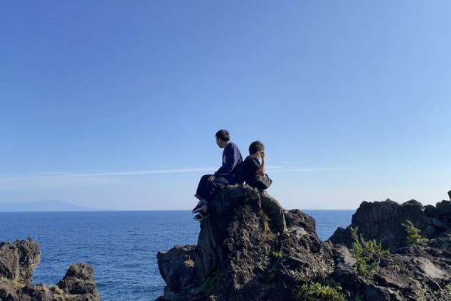 晴天のもと、海沿いの断崖絶壁に座って景色を眺める若者たちの画像。