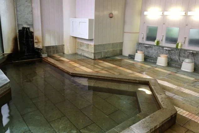 寮または従業員用の共同浴場（大浴場）。石造りの床と浴槽、洗い場にはシャワーと鏡、白い桶が並び、浴槽には湯が張られている。