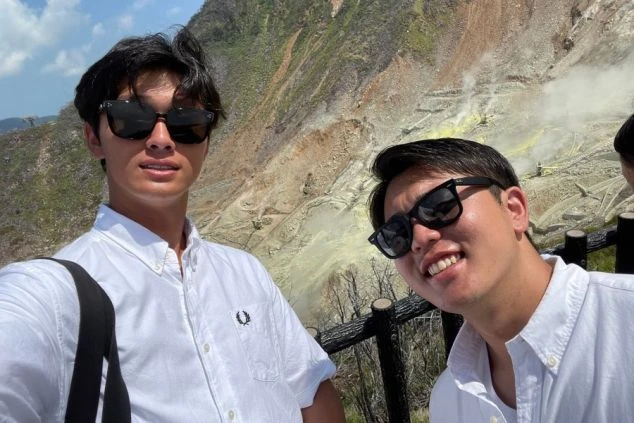 白いシャツにサングラスをかけた2人の男性。背景には白い噴煙が上がる荒々しい岩肌の山が広がっている。