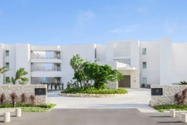 青空の下に建つ、白壁のモダンな低層ホテル。エントランス前には円形のロータリーがあり、中央に緑の樹木が植えられている。