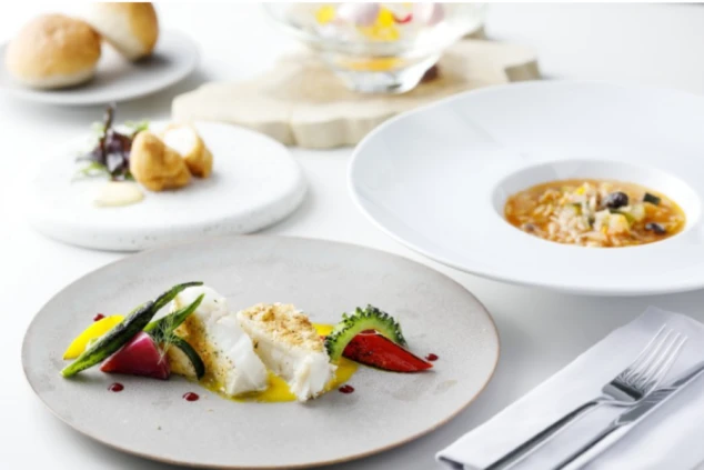 白い皿に美しく盛り付けられた魚料理。色鮮やかな夏野菜が添えられ、背景にはスープやパンなどのコース料理が並んでいる。